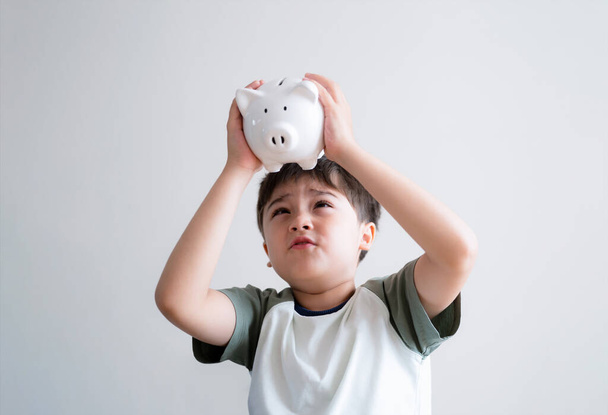 Emotional portrait Kid boy with empty piggy bank on white background, Isolated Mixed race Child looking at underneath money saving box.School kid Learning about planning saving money for future - Photo, Image