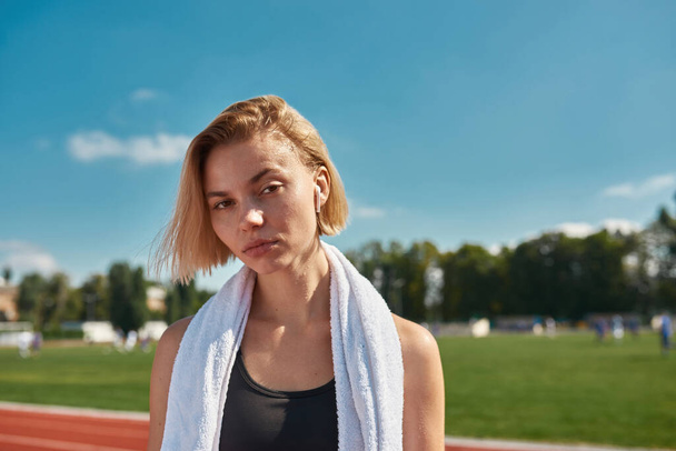 Close up portrait of serious sporty young woman with towel around her neck looking at camera while posing on the track at stadium on a sunny day - Foto, afbeelding