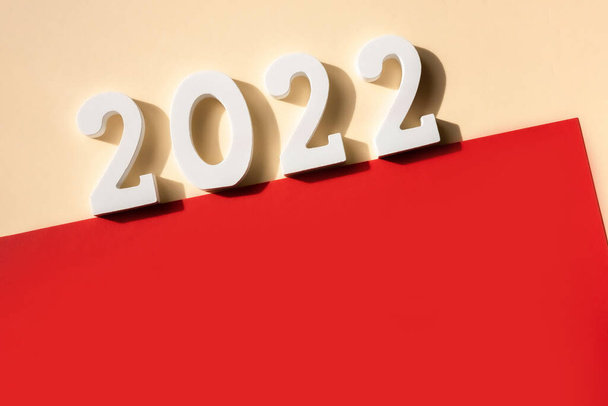Новый 2022 год. Белый номер 2022 и чистая красная бумага с копировальным пространством на бежевом фоне. Рождественская открытка. Чистый лист бумаги. Шаблон приглашения. Вид сверху, плоский, копировальное пространство. - Фото, изображение