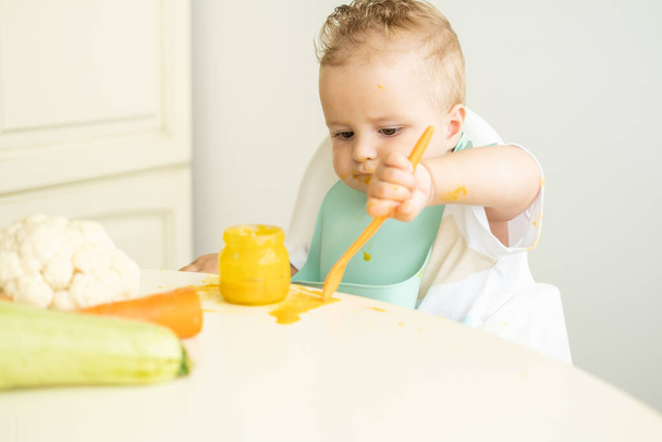 子供の椅子に座ってスプーンで野菜のピューレを食べる二人組の面白い赤ちゃん。子供は自分で食べることを学ぶ. - 写真・画像