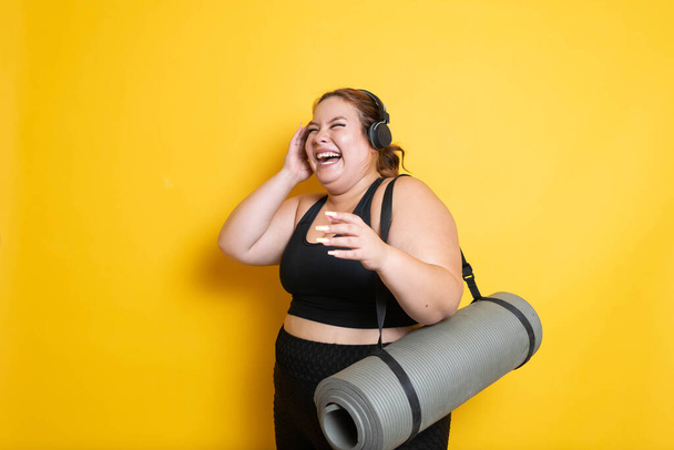 Молодая плюс размер фитнес-женщина в спортивной одежде слушать музыку на наушниках и смеяться вслух с ковриком йоги - Фото, изображение