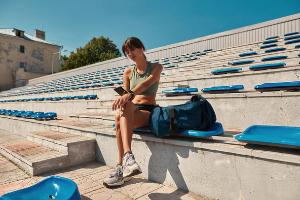Full length shot of fit young woman using smartphone, scrolling social media feed while resting after exercising, sitting on the stadium seat outdoors - Foto, Bild