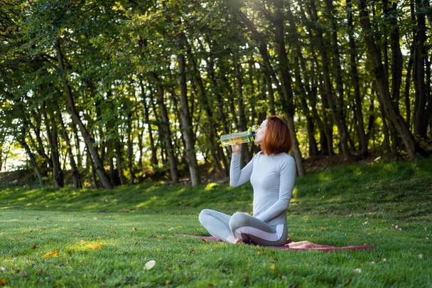 Spor elbiseli, spor elbiseli, spor yaparken su içen, sonbahar güneşli bir günde parkta yoga yapan bir kadın.. - Fotoğraf, Görsel
