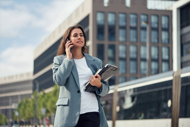 Beautiful formal dressed business woman in suit with a laptop and notebook outdoor. Brunette caucasian female, standing near office. - Foto, Imagen