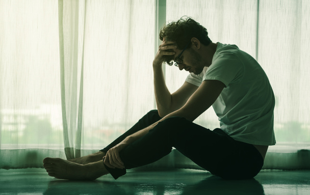 Καυκάσιος άντρας κάθεται στο πάτωμα δίπλα στα παράθυρα με καταθλιπτικά συναισθήματα για προβλήματα και προβλήματα.. - Φωτογραφία, εικόνα
