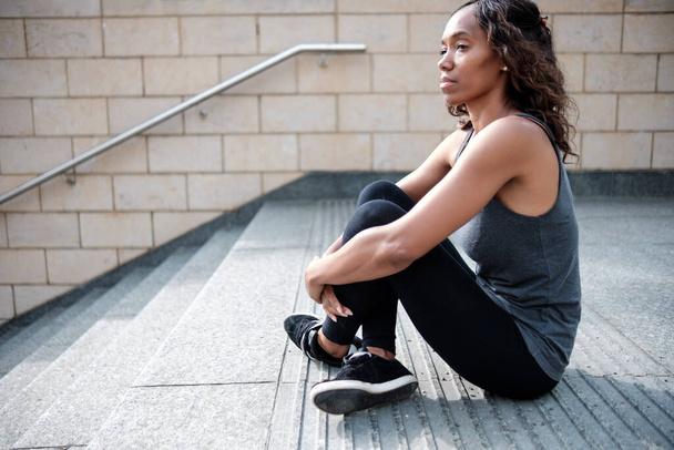 Ώριμη μαύρη γυναίκα που χαλαρώνει μετά την άσκηση. Φοράει ενεργά ρούχα και είναι σε επιχειρηματικό περιβάλλον.. - Φωτογραφία, εικόνα