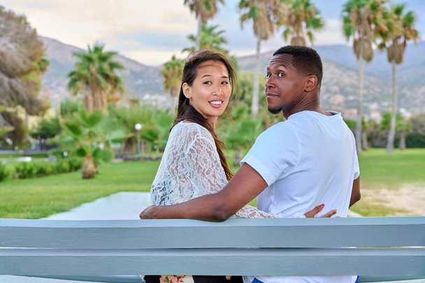 Tropikal bir parkta bankta oturan çok ırklı güzel bir çift. - Fotoğraf, Görsel