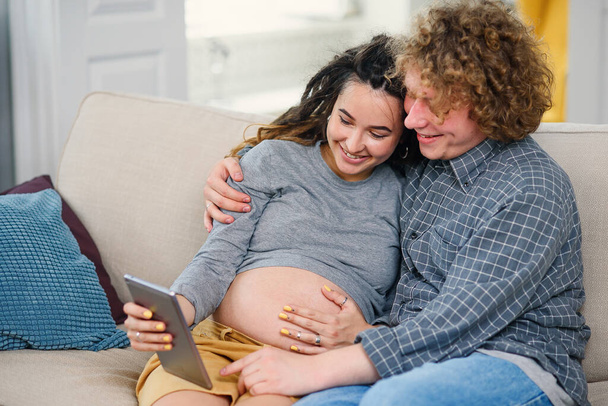 Good-looking pregnant couple relaxing on the comfortable couch at home and watching tv shows on tablet pc. - Fotó, kép