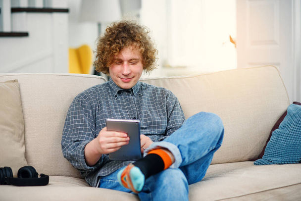 Young hipster man with curly hair sitting on the sofa and holding tablet in hands. - Fotó, kép