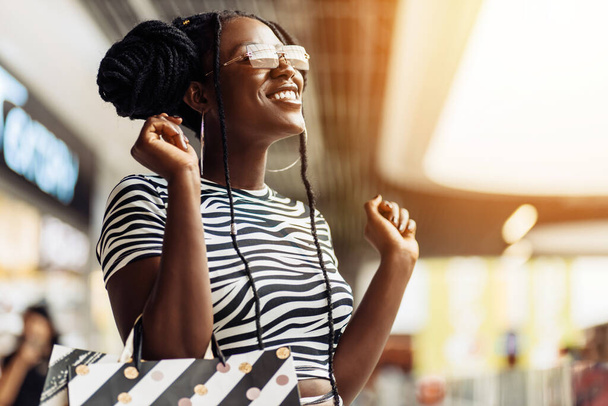 Portrait of smiling young black woman, wearing glasses, holding shopping bags, shopping mall, shopping concept, consumerism - Фото, зображення