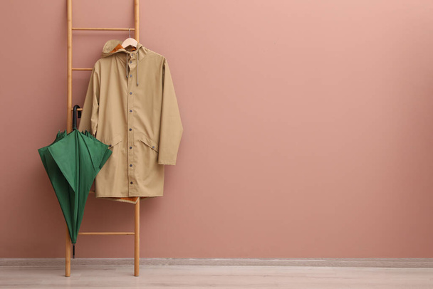 Stylish green umbrella and raincoat on wooden ladder near beige wall indoors. Space for text - Fotografie, Obrázek