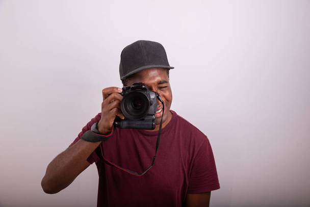 El fotógrafo toma una foto mientras mira a través del visor de la cámara. Fotógrafo africano aislado sobre fondo blanco. Fotografía tomada en el estudio. Concepto de fotógrafo profesional - Foto, imagen