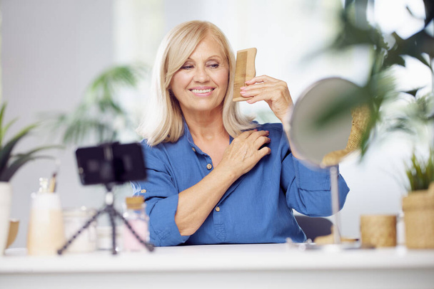 A happy blond senior woman is sitting at home and combing her hair. Daily routine - Photo, Image