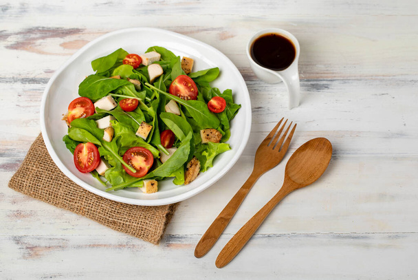 Fresh spinach salad with tomato, small pieces of herb roasted chicken in white ceramic dish on brown sackcloth, together with balsamic vinegar and utensil on white wooded table. Concept for healthy. - Photo, Image