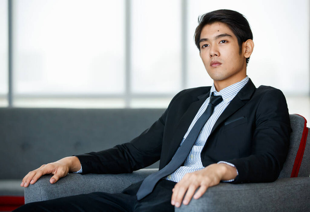 Muotokuva 20s komea nuori menestyvä aasialainen liikemies yllään muodollinen puku solmio, hymyillen luottavaisesti ja älykkäästi, istuu mukava sohva sisätiloissa toimisto. - Valokuva, kuva