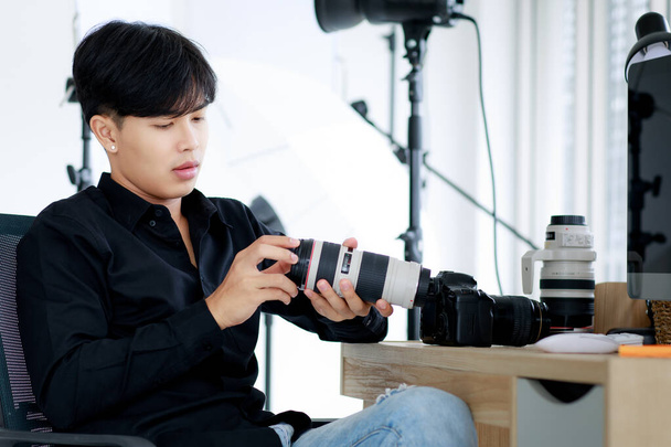 Asijský pohledný muž fotograf nosí ležérní černé tričko, drží fotoaparát, pózuje a sedí ve studiu se světelným vybavením - Fotografie, Obrázek