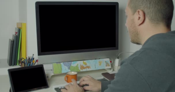 Retrovisione di un uomo caucasico con i capelli corti durante una sessione di lavoro sul computer desktop e un tablet in ufficio - Filmati, video