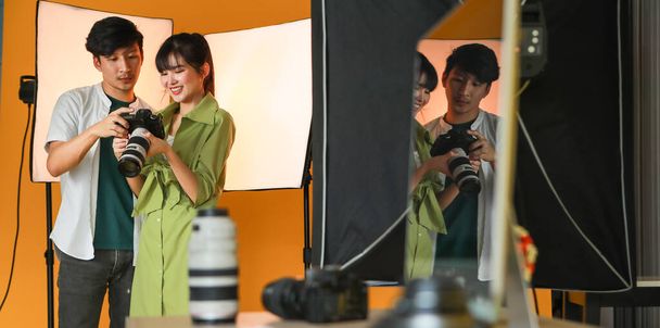 Bella giovane asiatica fotografa femminile che mostra un'immagine a un uomo modello e si sente molto soddisfatto dopo aver scattato la sua foto con la fotocamera digitale in un moderno studio luminoso - Foto, immagini