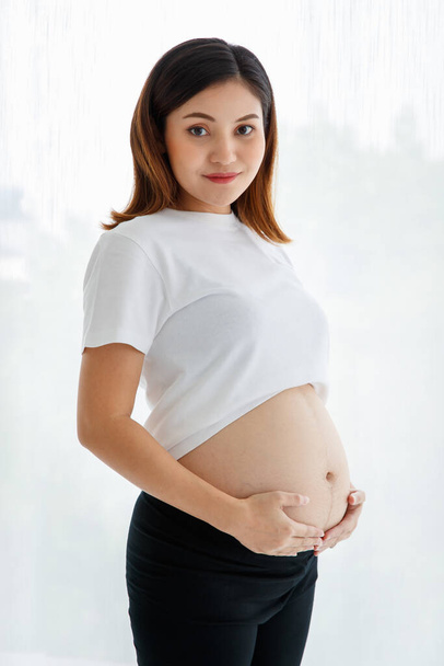 Smiley junge glückliche schwangere Asiatin trägt lässig stehend und berührt ihren Bauch mit Liebe und Sorgfalt Blick auf Kamera für Studio-Aufnahme vor weißem Hintergrund. - Foto, Bild
