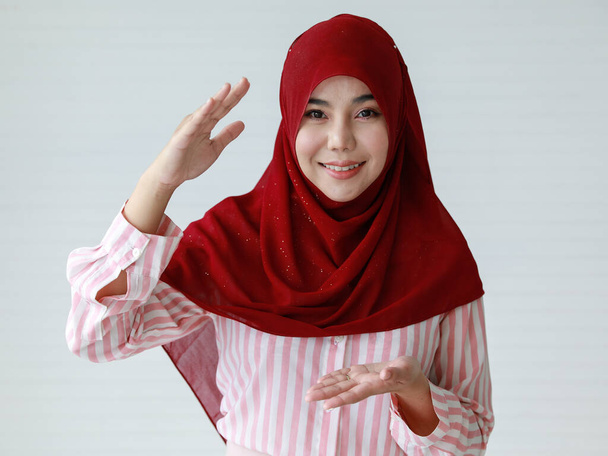 Fröhliche junge ethnische Frauen in lässiger Kleidung und traditionellem muslimischen Kopftuch lächeln und heben die Hand, um etwas vor weißem Hintergrund zu erklären - Foto, Bild