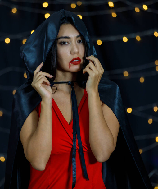 Mladý asijský upír žena v halloween oblečení s černou kapuci na hlavě a červené rty s kapající krví při pohledu na kameru proti tmavému pozadí s rozmazanými světly - Fotografie, Obrázek