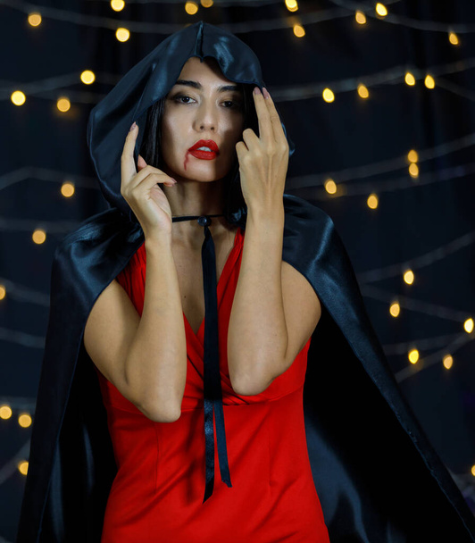 Młoda Azjatka wampirzyca w halloweenowym stroju z czarnym kapturem na głowie i czerwonymi ustami z kapiącą krwią patrząca w kamerę na ciemne tło z rozmytymi światłami - Zdjęcie, obraz