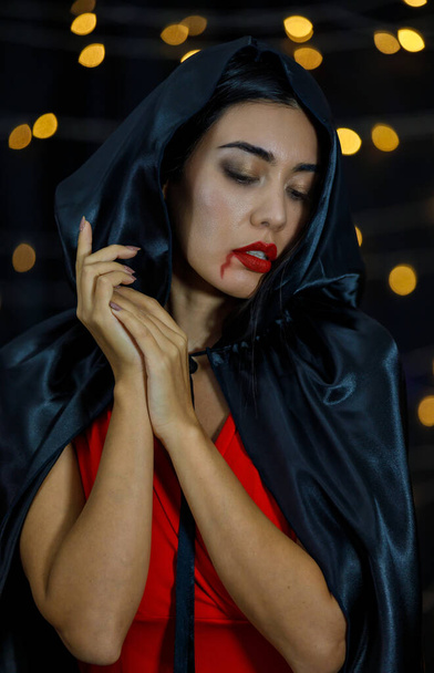 Mladá tajemná etnická žena s krví na červených rtech v černém plášti s kápí, dotýkající se brady a dívající se do kamery - Fotografie, Obrázek