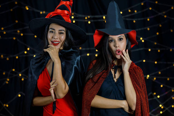 Affascinanti giovani ragazze in mantelli da strega e cappelli con le labbra rosse guardando la fotocamera mentre celebrano Halloween insieme durante la festa notturna - Foto, immagini