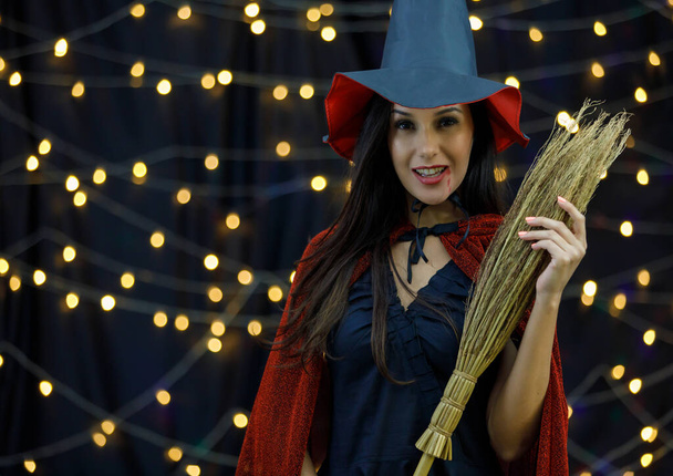 Zijaanzicht van charmante jonge dame met heksenhoed en cape paardrijden bezem en kijken naar de camera tijdens de nacht Halloween viering - Foto, afbeelding