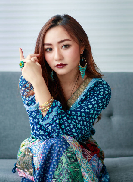 Joven, hermosa chica asiática linda, vestida en estilo gitano, ropa casual con confianza en sí misma manera, sentado en el sofá y mirando a la cámara. - Foto, imagen