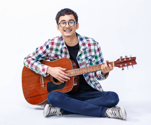 Πορτρέτο ενός χαριτωμένου χαμογελαστού νεαρού αγοριού που κρατάει την ακουστική κιθάρα. Επαγγελματίας κατώτερος κιθαρίστας κάθεται και παίζει ένα όργανο ενώ κοιτάζει κάμερα απομονωμένο λευκό φόντο - Φωτογραφία, εικόνα