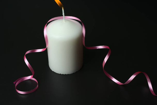 A large white burning candle on a black background with a pink ribbon. - Zdjęcie, obraz