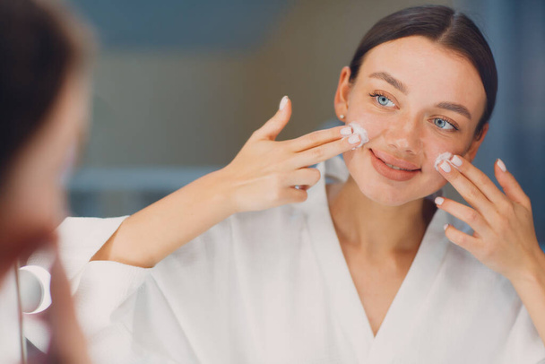 jonge vrouw aanbrengen cosmetische witte crème op haar gezicht bij spiegel - Foto, afbeelding