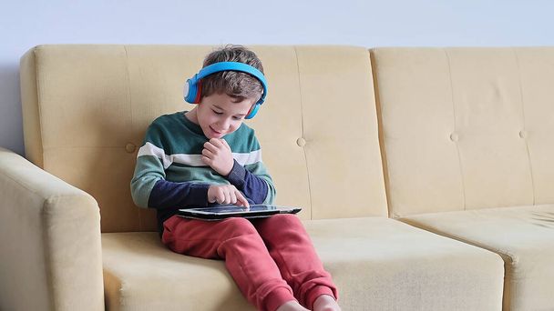 Veselý chlapeček se usmívá, zatímco sedí na gauči a doma používá tablet. Moderní dětská a vzdělávací technika. malý chlapec při pohledu na dotykovou podložku doma. - Fotografie, Obrázek