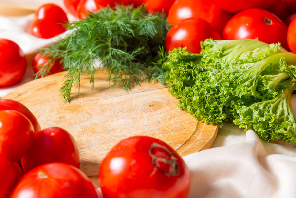 Tomaten, Dill und Salat liegen auf einer Tischdecke um ein Holzschneidebrett herum.. - Foto, Bild