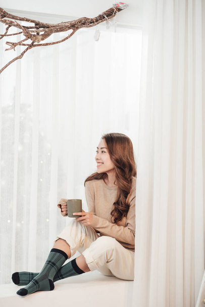Χαμογελώντας όμορφη γυναίκα κάθεται στο σπίτι με φλιτζάνι καφέ πάνω από τα φώτα φόντο.  - Φωτογραφία, εικόνα