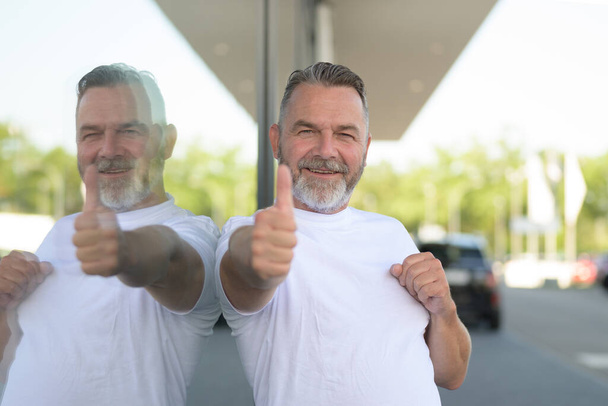 Homme âgé souriant enthousiaste donnant un geste de pouce vers le haut du succès avec un sourire rayonnant reflété dans le verre d'une grande fenêtre de magasin en ville - Photo, image