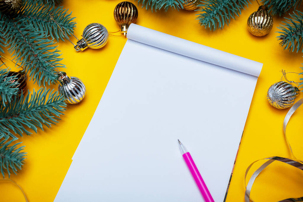 Σημειωματάριο με αντίγραφο χώρου με ροζ στυλό σε κίτρινο φόντο. Χριστουγεννιάτικο φόντο με σημειωματάριο για σημειώσεις, κλαδιά ερυθρελάτης και χριστουγεννιάτικα παιχνίδια - Φωτογραφία, εικόνα