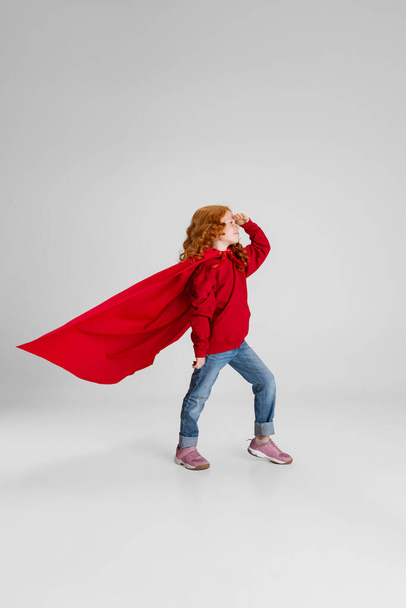 full-length back side view retrato de pequena menina encaracolada, criança em manto vermelho apontando em algum lugar isolado sobre fundo estúdio cinza. - Foto, Imagem