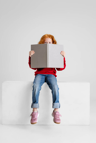 Full-length portrait of little curly girl, child sitting with book covering face isolated over gray studio background. - Foto, Imagen