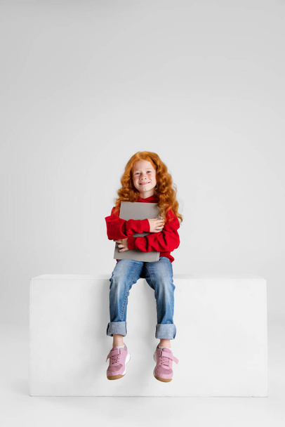 Full-length portrait of little curly smiling girl, child sitting with tablet isolated over gray studio background. - Foto, Imagem