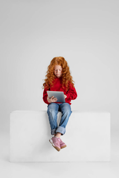 Full-length portrait of little curly smiling girl, child sitting and typing on tablet isolated over gray studio background. - Foto, imagen