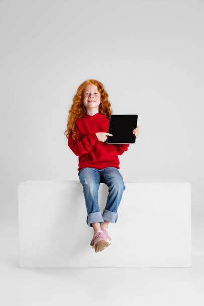 Portrait of little red-haired curly girl, child pointing at tablet isolated over gray studio background. - Фото, зображення