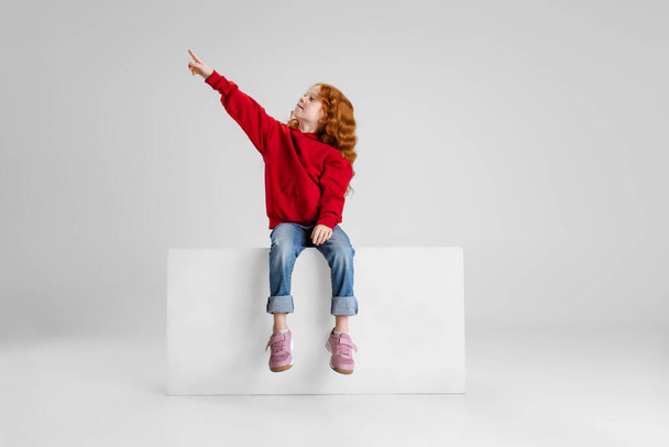 Портрет маленькой рыжеволосой кудряшки, ребенка, указывающего где-то в изоляции на сером фоне студии. - Фото, изображение