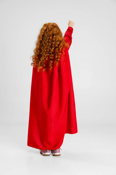 Full-length back view portrait of Caucasian little curly redheaded girl pointing somewhere isolated over gray studio background. - Foto, imagen