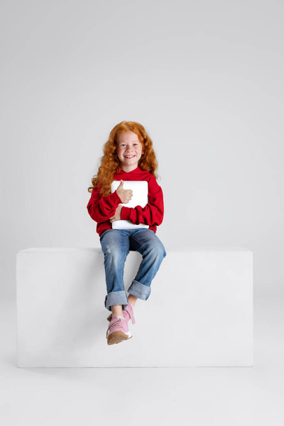 Портрет маленькой улыбающейся девочки, ребенка с планшетом, позирующего изолированно на сером фоне студии. Счастливого детства - Фото, изображение