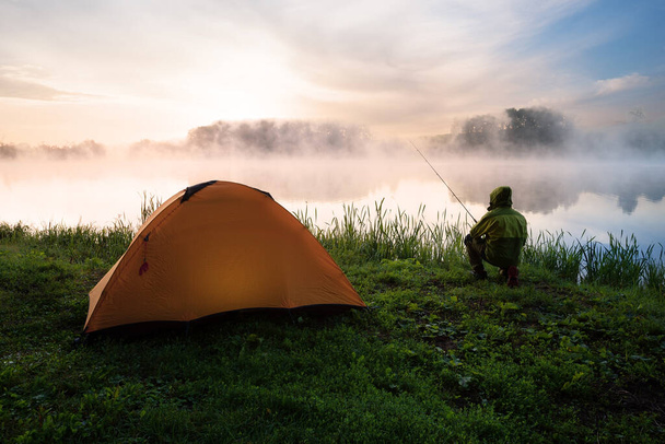 Pescador sentado na costa do lago nebuloso perto de uma tenda laranja - Foto, Imagem