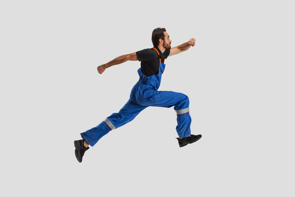 Portrait of young man, male auto mechanic in dungarees running isolated over white studio background. Concept of labor, occupation, funny meme emotions - Φωτογραφία, εικόνα