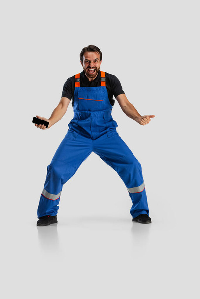 One young excited man, male auto mechanic or fitter in dungarees using phone isolated over white studio background. Concept of occupation, career, funny meme emotions - Photo, Image