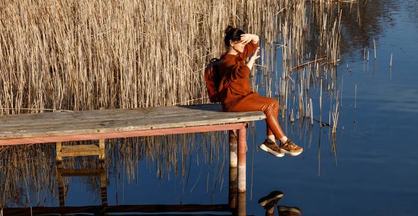 Porträt einer Frau in herbstlicher orangefarbener Kleidung mit Rucksack, die auf einer Holzbrücke in der Nähe des Sees mit blauem Wasser sitzt und in die Sonne blickt. Menschen, Reisen, Naturkonzept. - Foto, Bild